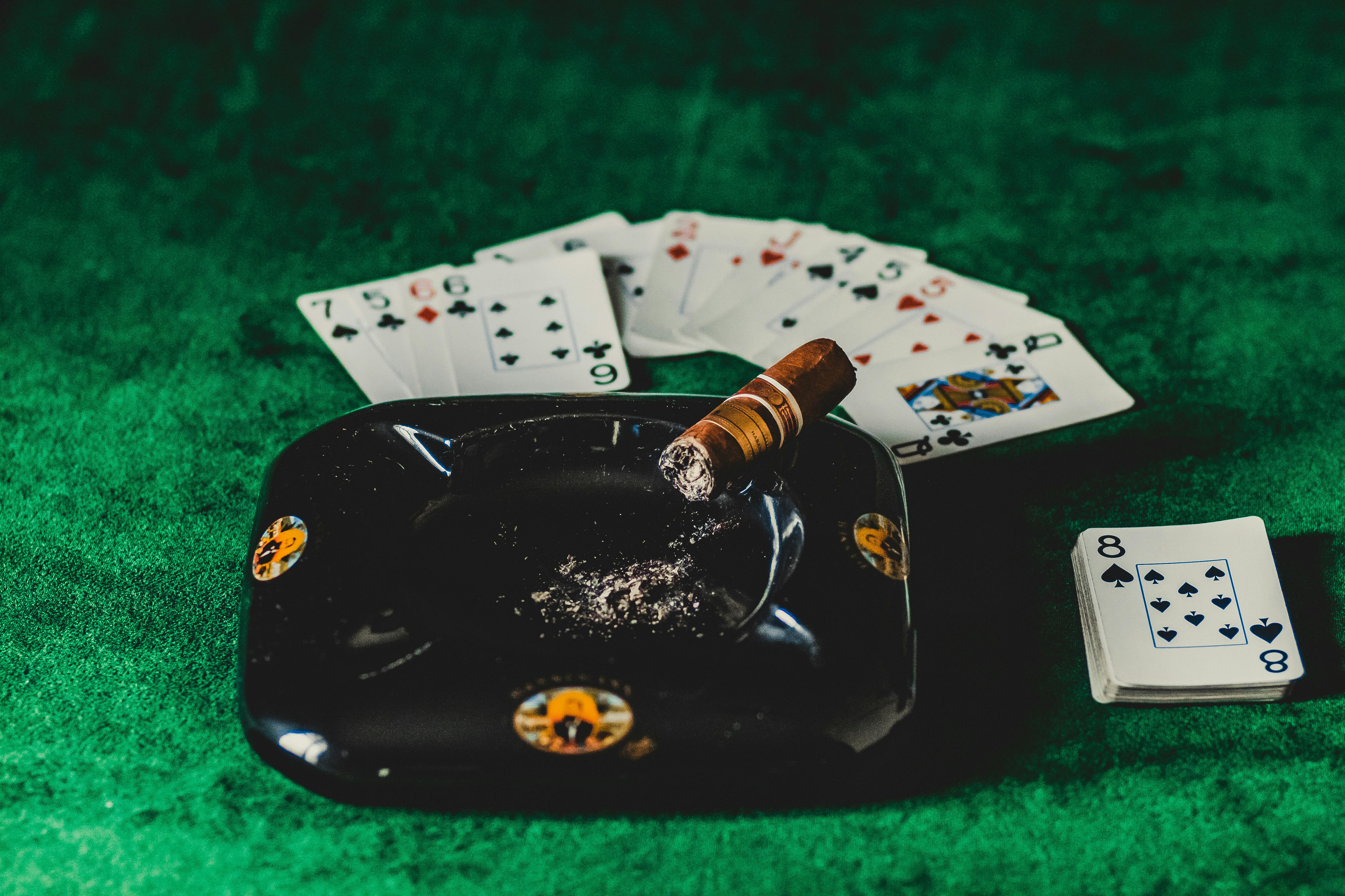 A poker table with playing cards, a lit cigar, and an ashtray on a green felt surface.